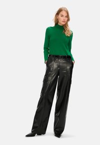 Grüner taillierter Rollkragenpullover kombiniert mit hoch taillierten, schwarzen Lederröhrenhosen mit weitem Bein, die Seitentaschen und ein elegantes Design aufweisen.