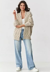 Jasno beżowy sweter z dzianiny o fakturowanym wzorze w warkocze, z otwartym przodem i długimi rękawami, w połączeniu z jasnoniebieskimi spodniami typu wide-leg i białymi trampkami.