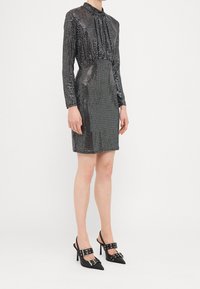 Robe noire à sequins avec manches longues et encolure froncée, associée à des escarpins noirs à bout pointu avec des brides décoratives à boucle.