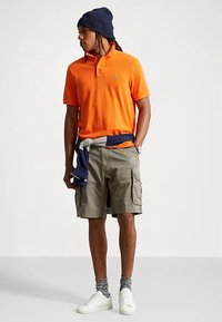 Oranžové polo tričko, khaki cargo šortky s bočnými vreckami, biele tenisky, sivé ponožky so vzorom, námornícka čiapka a modrý sveter priviazaný okolo pása.