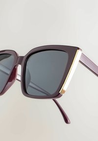 Bordové slnečné okuliare s veľkými uhlovými šošovkami, zlatým akcentom na vonkajšom ráme a hladkou, lesklou textúrou. Rám pokračuje do rovnomerných ramienok.