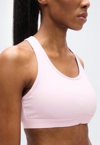 Αθλητικό σουτιέν σε ανοιχτό ροζ χρώμα από απαλό, ελαστικό ύφασμα. Διαθέτει σχέδιο racerback και μικρό λευκό λογότυπο στο μπροστινό μέρος.