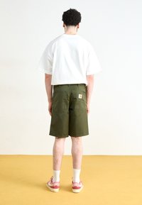 Tricou alb supradimensionat și pantaloni scurți verde măsliniu cu două buzunare la spate. Pantalonii scurți au un patch cu logo-ul unei mărci. Încălțămintea este formată din adidași roz și alb.