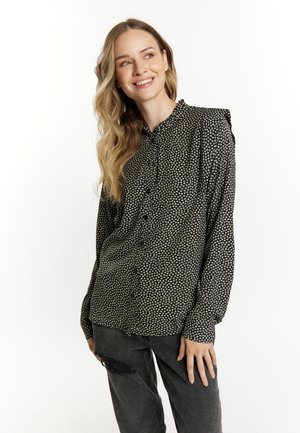 Frau mit langen welligen Haaren, die eine schwarze gemusterte Bluse und dunkle zerrissene Jeans trägt, lächelt und schaut seitlich vor einem weißen Hintergrund.