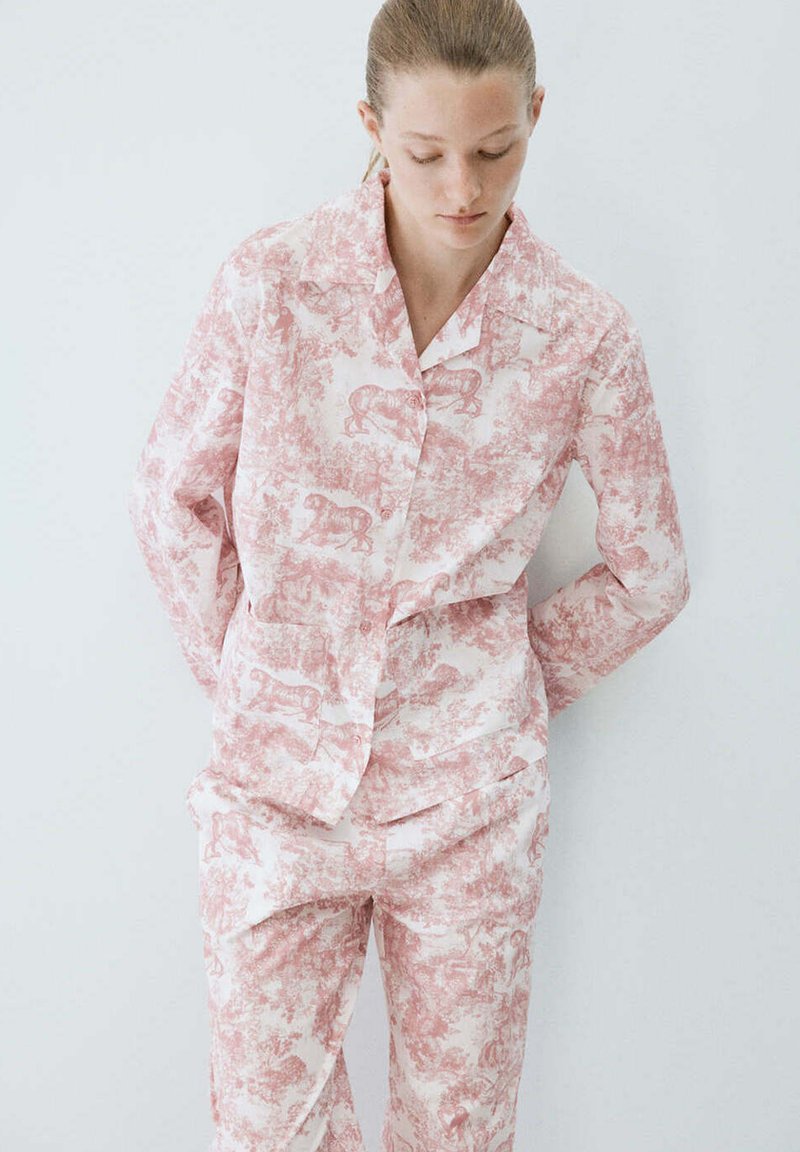 Femme portant une chemise à manches longues et un pantalon assortis rose et blanc à motifs d’animaux et de nature, debout devant un fond blanc uni.