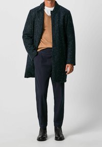 Mörkaspräcklig kappa med krage, knappad framtill, tillsammans med en brun tröja och marinblå byxor. Svarta ankelstövlar fulländar looken.