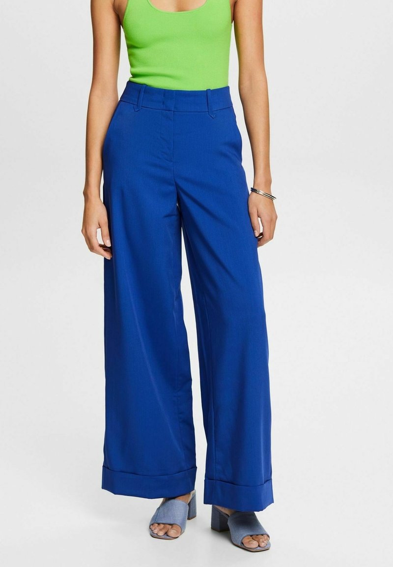 Femme portant un pantalon large bleu vif, un top vert sans manches, des sandales à talons bleu clair et un bracelet argenté au poignet droit.
