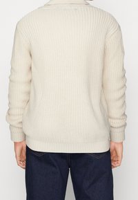 Pletený svetr v krémové barvě s texturovaným vzorem, dlouhými rukávy a žebrovanými manžetami a lemem, stylizovaný přes tmavé džínové kalhoty.