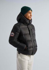 Schwarze Daunenjacke mit hohem Kragen und vorne Reißverschluss, mit einem Logo-Patch am linken Arm. Trägt eine grüne Strickmütze und hellfarbige Hosen.