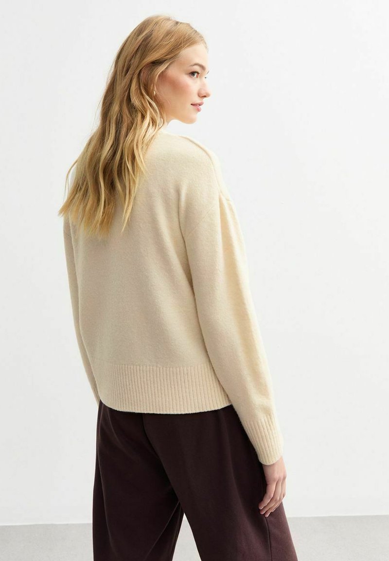 Beiger Pullover mit geripptem Saum, oversized Passform und überschnittenen Schultern, kombiniert mit dunkelbraunen, weit geschnittenen Hosen. Sanfte Textur, minimales Design.