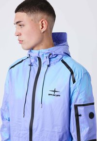 Giovane con capelli corti che indossa una giacca a cappuccio viola chiaro e blu con zip nera e logo PXP, rivolto a sinistra.