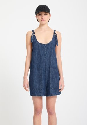 Jeune femme portant une combishort en denim bleu foncé sans manches avec des liens aux épaules et une casquette noire Volcom, debout devant un fond blanc uni.