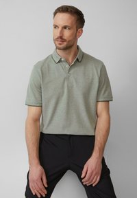 Hellgrünes Poloshirt aus strukturiertem Stoff, mit einem Kragen mit kontrastierenden Besätzen und drei Knöpfen. Getragen mit schwarzen Hosen.