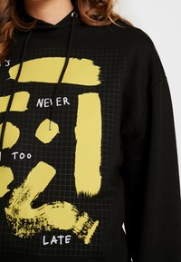 Sudadera negra con gráficos de pinceladas amarillas en negrita y el texto en blanco "NUNCA ES DEMASIADO TARDE" sobre un fondo con patrón de cuadrícula. Material de algodón, ajuste relajado.