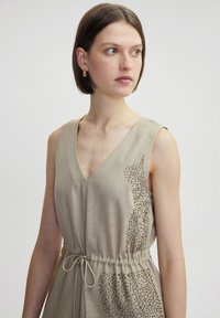 Abito beige senza maniche con scollatura a V, vita con cordoncino e motivo punteggiato decorato sul lato, caratterizzato da una morbida texture simile al lino.