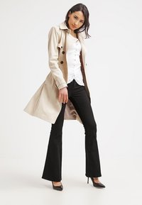 Beige trenchcoat met dubbele knopen, witte blouse met kantafwerking, zwarte flared broek en zwarte hoge hakken.