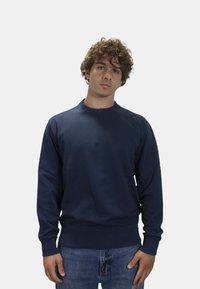 Felpa girocollo navy in un tessuto morbido con polsini e orlo a coste; dettaglio del logo sul colletto. Indossata con jeans in denim blu.
