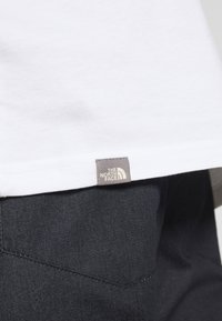 Vitt klädesplagg med en liten grå etikett märkta "The North Face" ovanför mörkblå byxor eller jeans.