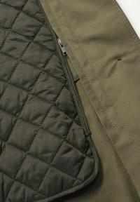 Veste olive en vert avec une doublure intérieure noire matelassée, une fermeture éclair, et une texture lisse avec des matériaux contrastants et des coutures détaillées.