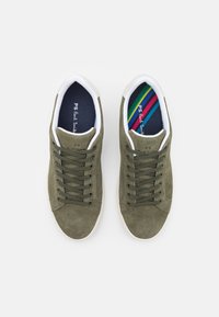 Olivgröna mockaskor med vita gummi-sulor, med mörkgröna snören och ett mönstrat, flerfärgat foder. Logotyp synlig på innersulan.