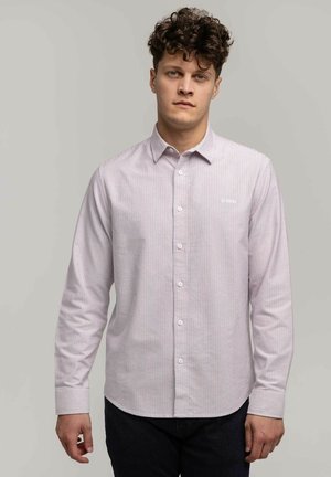 Młody mężczyzna z kręconymi włosami, ubrany w jasnoróżową koszulę w białe paski z guzikami i ciemne spodnie, stojący na jednolitym szarym tle.