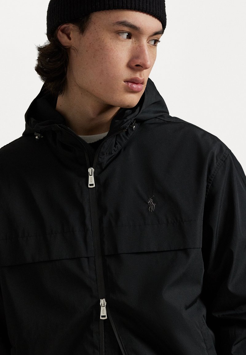 Veste noire à capuche avec zip frontal, grandes poches, et logo brodé. Texture lisse et design minimaliste, accompagnée d'un bonnet en tricot.