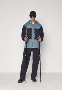 Windbreaker jacket in light blue and black with zippered pockets, paired with dark trousers and hiking poles. Features a hood and sunglasses.