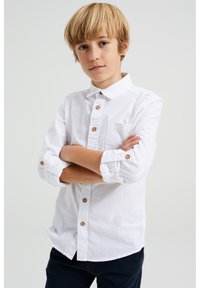 Bílá košile na knoflíky s ohrnutými rukávy, hnědými knoflíky, náprsní kapsou a hladkým povrchem, která je doplněna tmavými kalhotami.