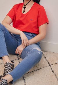 Rød kortærmet V-hals T-shirt med Levis logo, kombineret med lyseblå, revnede skinny jeans og mønstrede sorte sandaler. Sidder på et tekstureret tæppe.