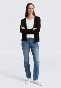 Črn lahkoten kardigan čez belo srajco, kombiniran z modrimi kavbojkami in belimi supergami. Kardigan ima sproščen kroj in odprt sprednji del.