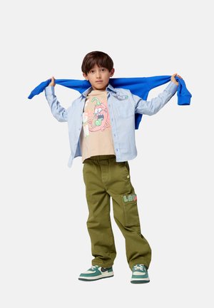 Lichtblauwe button-up overhemd, beige graphic t-shirt met kleurrijk ontwerp, olijfgroene broek en groene sneakers met zwarte accenten en witte zolen.