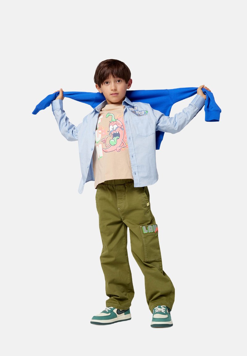 Chemise bleu clair à manches longues, t-shirt graphique beige avec un design coloré, pantalon vert olive et baskets vertes avec des accents noirs et des semelles blanches.