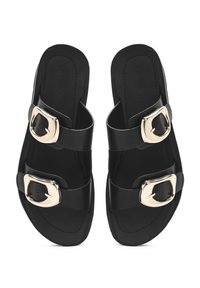 Sandalias planas negras con dos correas ajustables que presentan hebillas en tono dorado. Textura suave mate, suela plana y diseño abierto en la parte delantera.