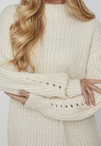 Pull en maille crème avec un col montant. Présente des poignets côtelés épais, des détails ajourés aux manches et une finition douce et texturée.