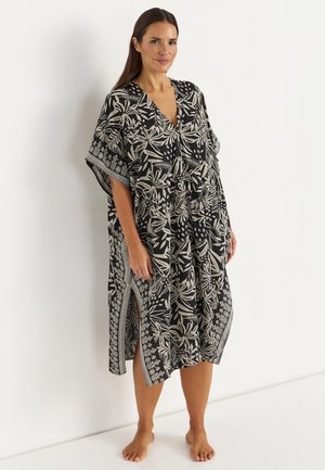 Kobieta stojąca boso, ubrana w luźną sukienkę w czarno-biały kwiatowy wzór z rozcięciami po bokach, na tle jednolitej białej ściany.