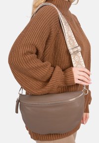 Braune Leder-Crossbody-Tasche mit runder Form, ausgestattet mit einem silbernen Reißverschluss und einem verstellbaren, gemusterten Riemen in Beige und Braun.