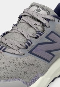 Grijze sportschoen met een gestructureerd mesh-bovenwerk, paarse accenten, prominent 'N' logo en patroonveters. Lichtgewicht constructie met gevoerde kraag.