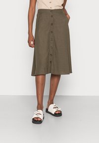 Gonna midi khaki realizzata in tessuto morbido con taglio dritto, dotata di chiusura frontale con bottoni e tasche laterali. Abbinata a sandali bianchi.