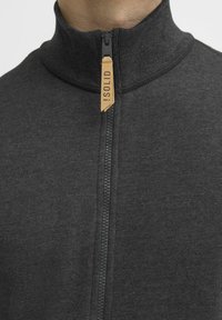 Sweat à zip gris foncé avec un col haut, présentant une fermeture éclair noire et une étiquette en cuir beige portant la mention « !SOLID. » Texture lisse sur l'ensemble.