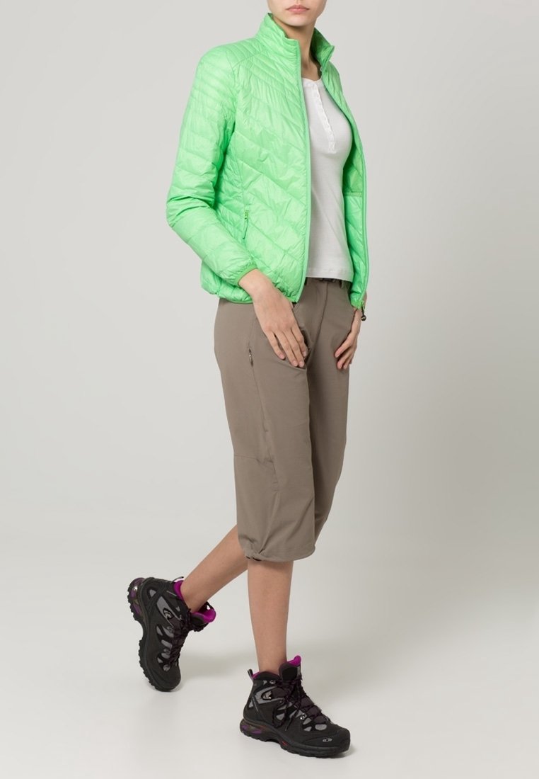 Femme portant une veste matelassée vert clair, une chemise blanche, un pantalon capri beige et des chaussures de randonnée noires avec des accents roses, debout sur un fond uni.