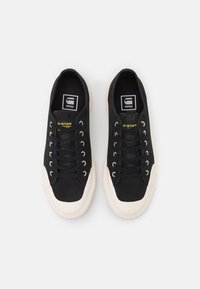 Zapatillas de lona negras con punta y suela de goma blanca. Presenta cordones negros, ojales y una etiqueta con el logo en la lengüeta.