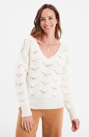 Weißer gestrickter Pullover mit V-Ausschnitt, offenem Muster und gerippten Bündchen. Kombiniert mit hellbraunen Hosen, die einen lässigen Look schaffen.