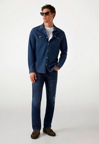 Jeansskjorta i mörkblått med två bröstfickor och knappstängning, kombinerad med mörkblå jeans och bruna mockasinliknande skor.
