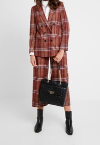 Kobieta ma na sobie dwurzędowy, rdzawo-szklany garnitur w kratę z szerokimi spodniami, białą koszulę, czarne skórzane botki za kostkę, trzyma czarną pikowaną torebkę na łańcuszkach w kolorze złota.