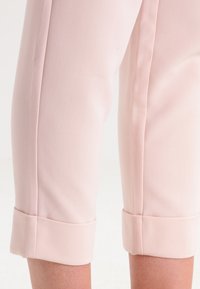 Zblízka na světle růžové kalhoty na míru, s manžetami ohrnutými těsně nad kotníky na nahých nohách.