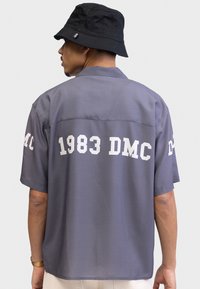 Siva majica kratkih rukava s bijelim natpisom "1983 DMC" na leđima i lijevom rukavu. Ima opušteni kroj i ovratnik; izrađena je od laganog materijala.