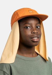 Persona joven que lleva una gorra de sol Buff naranja con solapa beige para el cuello y camiseta verde de cuello redondo, mirando ligeramente hacia la derecha con expresión neutral.