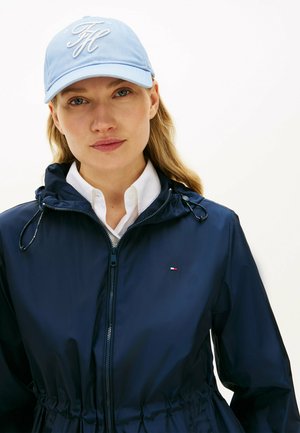 Kobieta z długimi blond włosami, ubrana w jasnoniebieską czapkę, białą koszulę z kołnierzykiem oraz granatową kurtkę na zamek z troczkami.