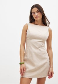 Une robe ajustée sans manches en lin beige clair, avec un col bateau et des coutures subtiles. Un bracelet vert est porté au poignet gauche.