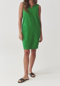 Green sleeveless dress with a v-neckline, knee-length cut, smooth texture, and minimalistic design. Worn with black slip-on sandals.